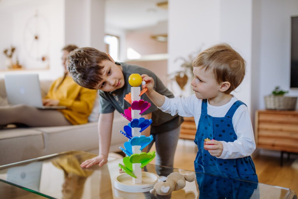 Little siblings playing with montessori wooden marble run in living room, their mother is sitting on